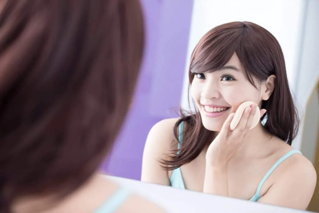 Smiling woman with brown hair and aqua shirt putting on makeup and smilling